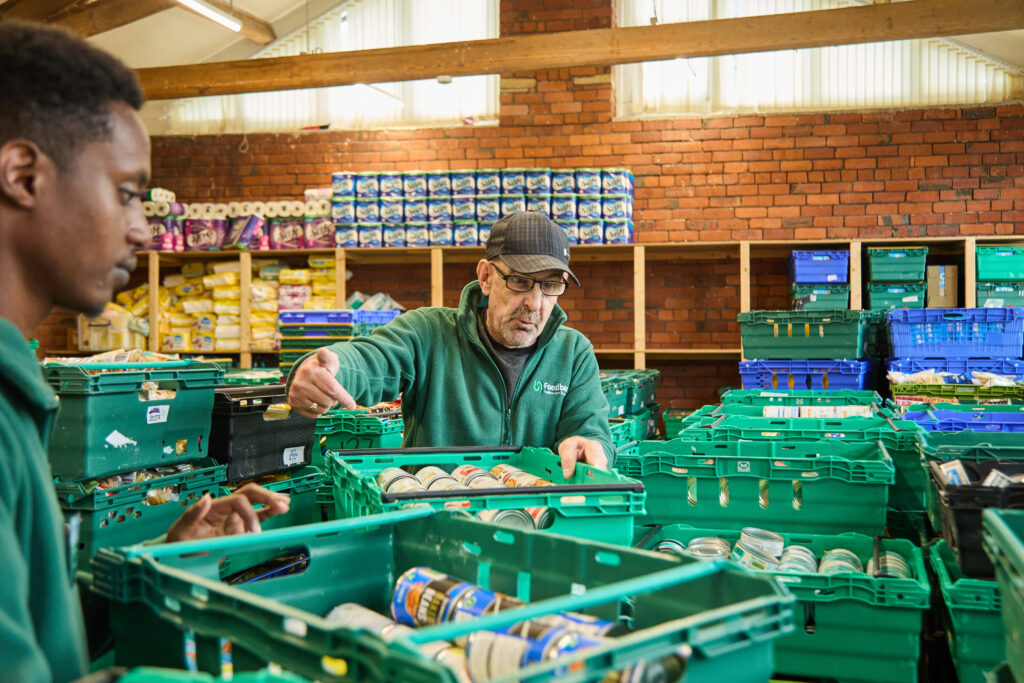 An older man and a younger man are looking over packed food crates, sorting food donations in a warehouse.